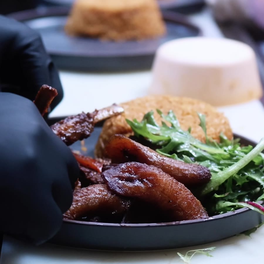 Plates of lamb, fried plantain and jollof rice being plated